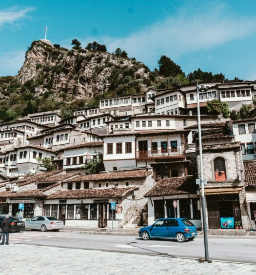 Uma rua de Berat, uma cidade da Albânia, com muitas casas com arquitetura antiga na cor branca, logo atrás delas, é possível ver muitas rochas com vegetação sob elas