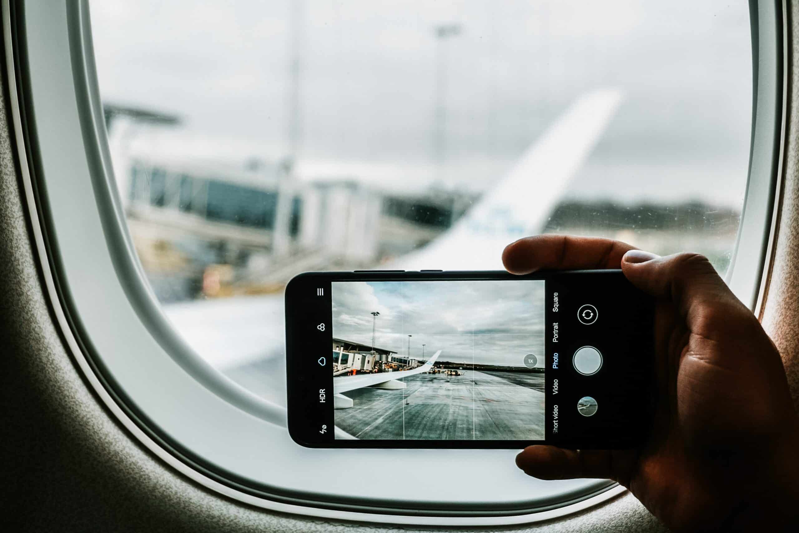 mão branca segurando um celular android na posição vertical para fotografar a asa de um avião vista de dentro da janela da aeronave