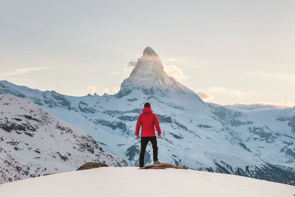 Um homem com um casaco vermelho e calças pretas virado de frente para uma enorme montanha cheia de neve