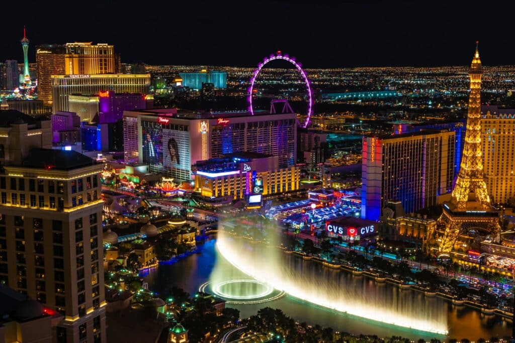Visão de Las Vegas Strip de noite, com muitas luzes coloridas acessas e atrações turísticas como a falsa London Eye e a Torre Eiffel, além de uma fonte grande toda iluminada e as ruas cheias