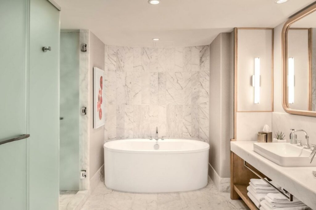 Banheiro do JW Marriott Parq Vancouver com uma banheira oval, chão de mármore, do lado esquerdo um box com portas de vidro temperado, do lado direito uma via larga e um espelho, na parte debaixo do móvel da pia há muitas toalhas brancas dobradas