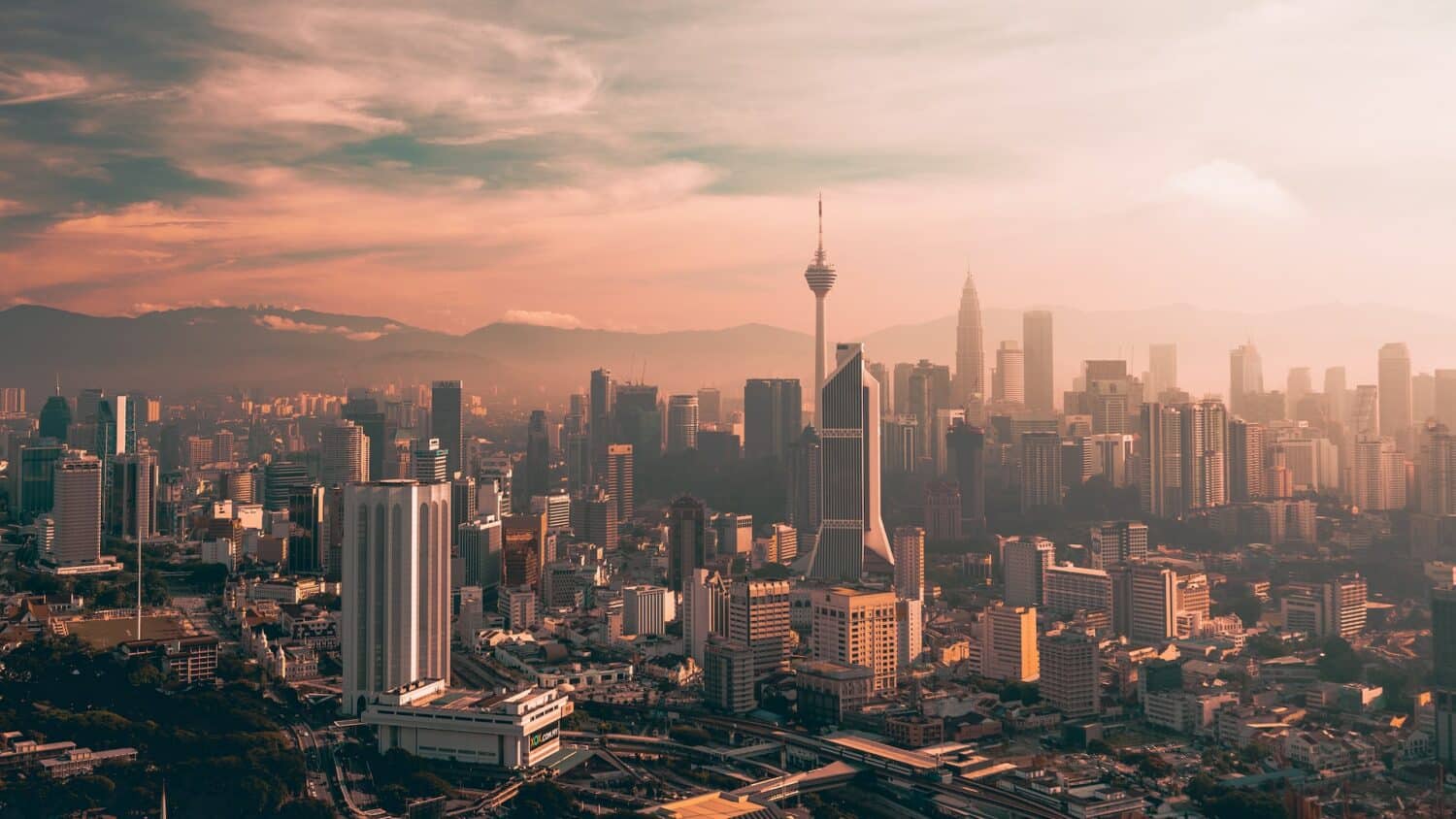 Centro de Kuala Lumpur visto de cima com prédios e arranha-céus modernos, estradas pavimentadas em um dia com céu nublado e alaranjado.