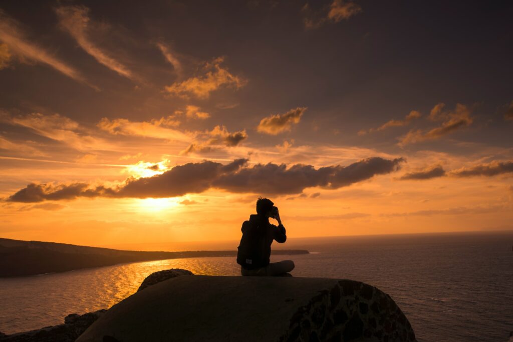 Um rapaz sentada no topo de uma rocha fotografando o mar de Santorini, a foto é escurecida porque é o pôr do sol, o céu está laranja com nuvens