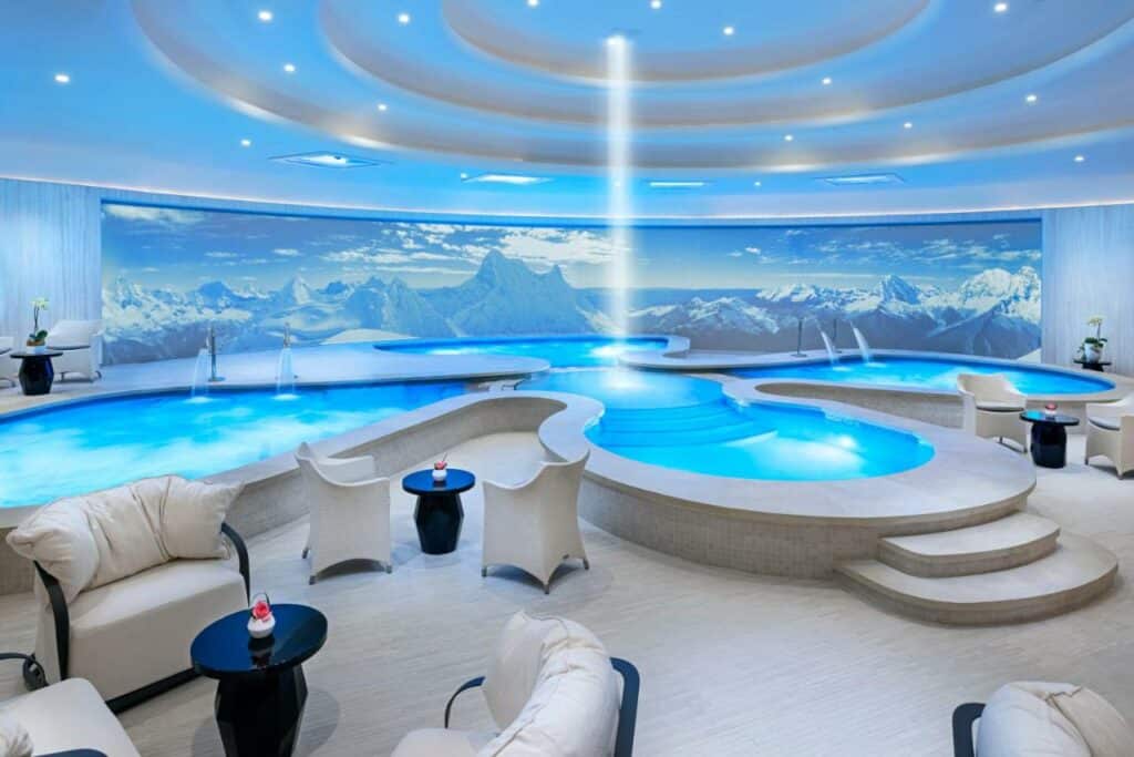 Piscina coberta do Las Vegas Hilton At Resorts World em formatos arredondados, iluminação indireta do teto, com poltronas e mesinhas ao redor, o deck é de madeira clara