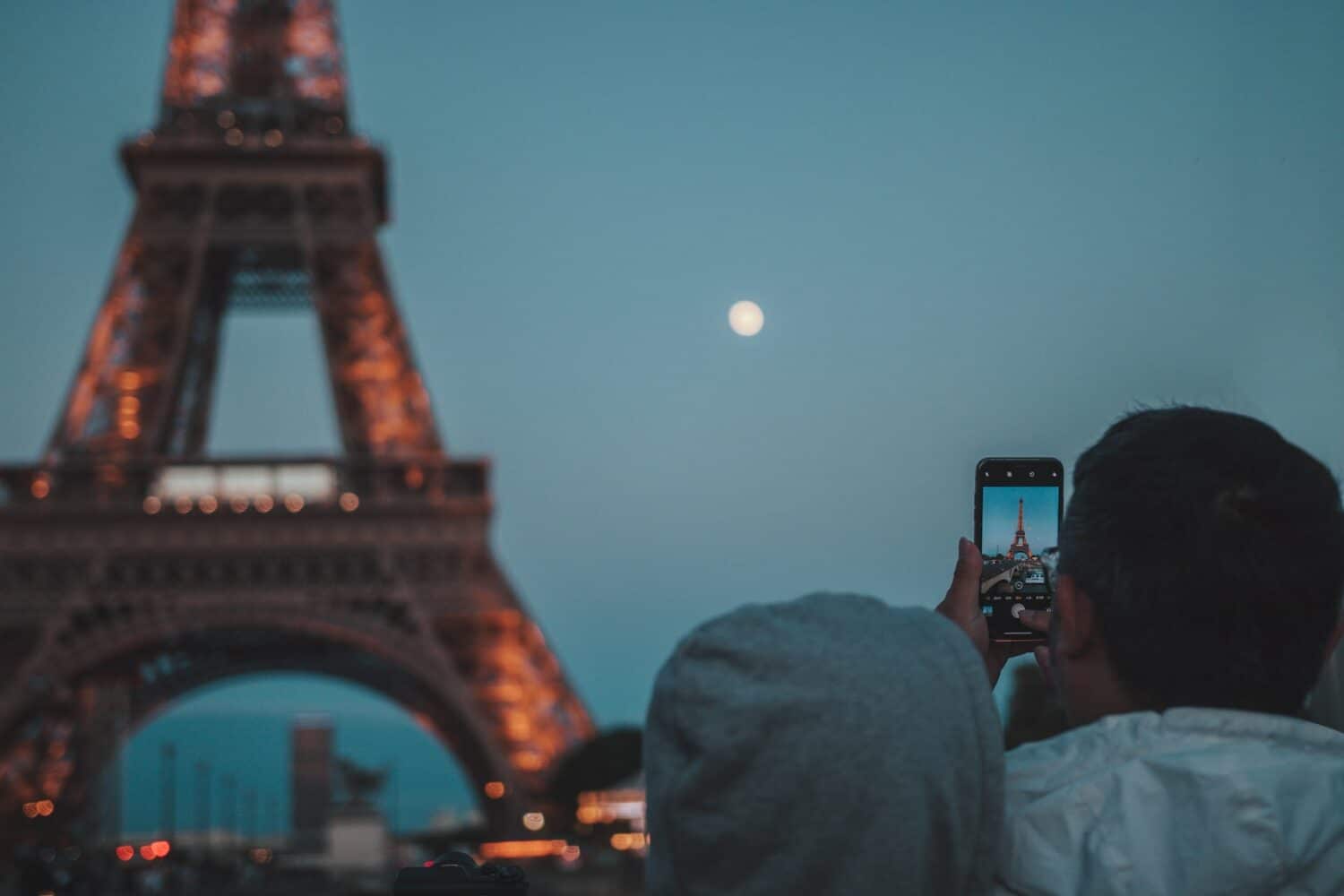 A Torre Eiffel iluminada de noite e sendo fotografada por um homem usando um celular