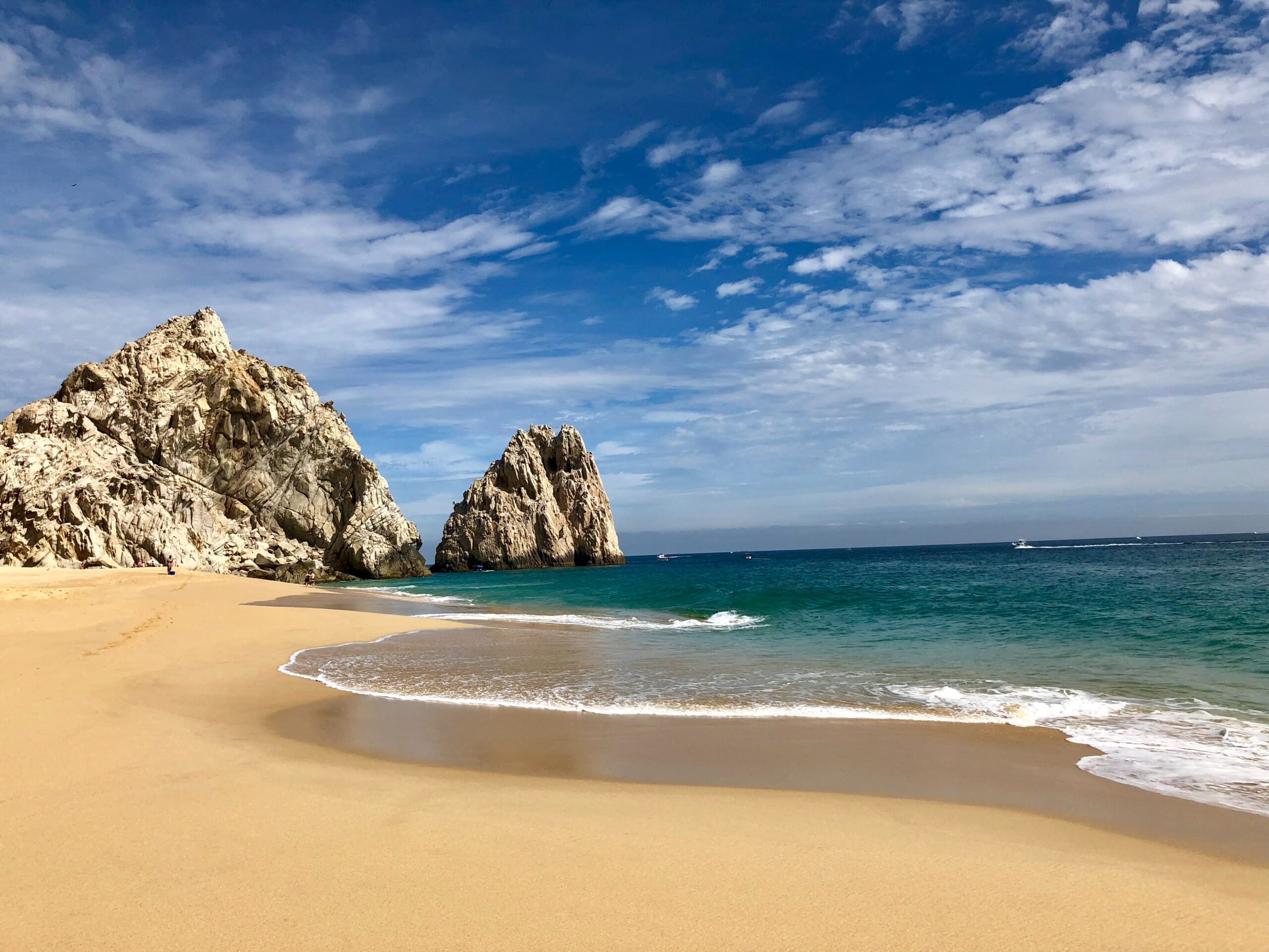 Praia Baja Califórnia em Los Cabos com areia, mar de tonalidade azul meio esverdeada, rochas perto da areia e no mar e céu azul.
