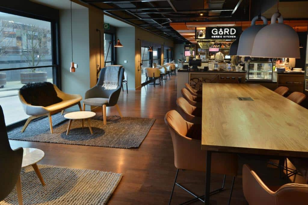 lounge com diversas poltronas noCourtyard by Marriott Brussels, um dos hotéis em Bruxelas, com o bar ao fundo e uma grande mesa de madeira no centro, com paredes de vidro