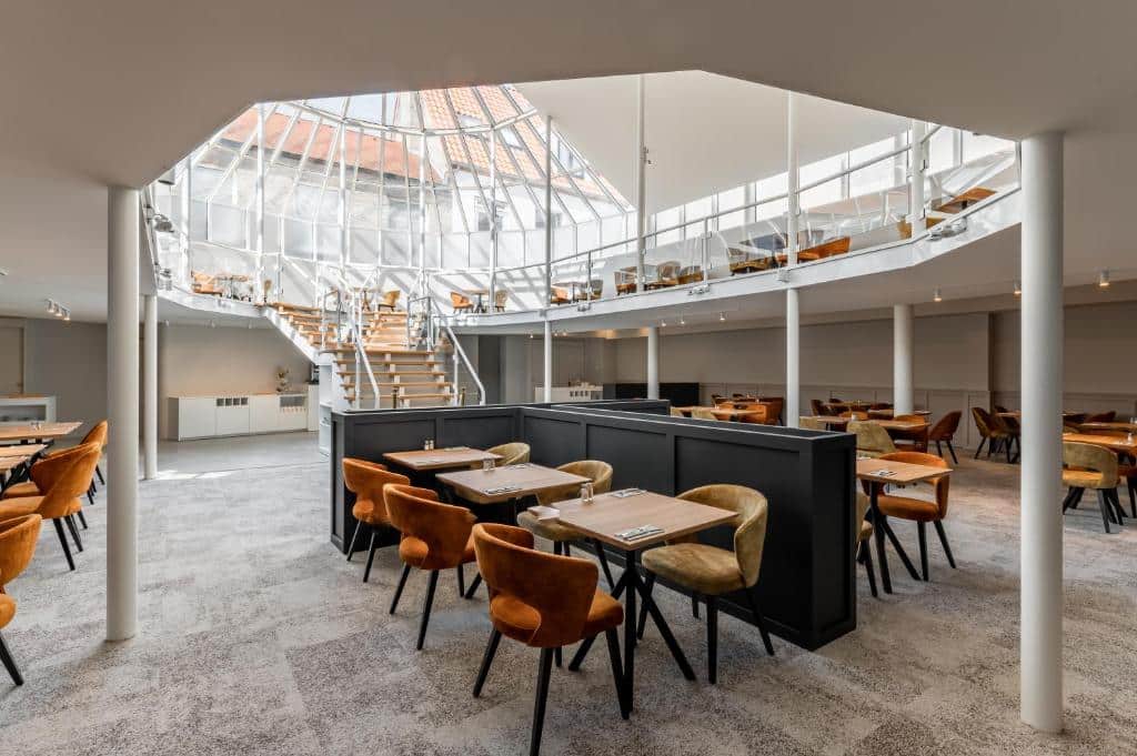 lounge do Hotel Le Bois De Bruges, com teto de vidro em uma abóbada pontuda e várias cadeiras cinza e alaranjadas com mesas de madeira abaixo em dois andares e um ambiente amplo
