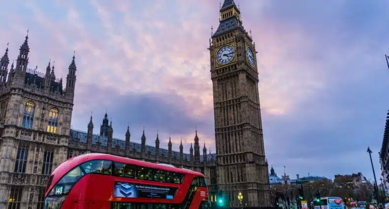 Rua movimentada em Londres com um dos icônicos ônibus vermelho passando, e ao fundo, dá para ver a torre do Big Ben em Londres, no Reino Unido