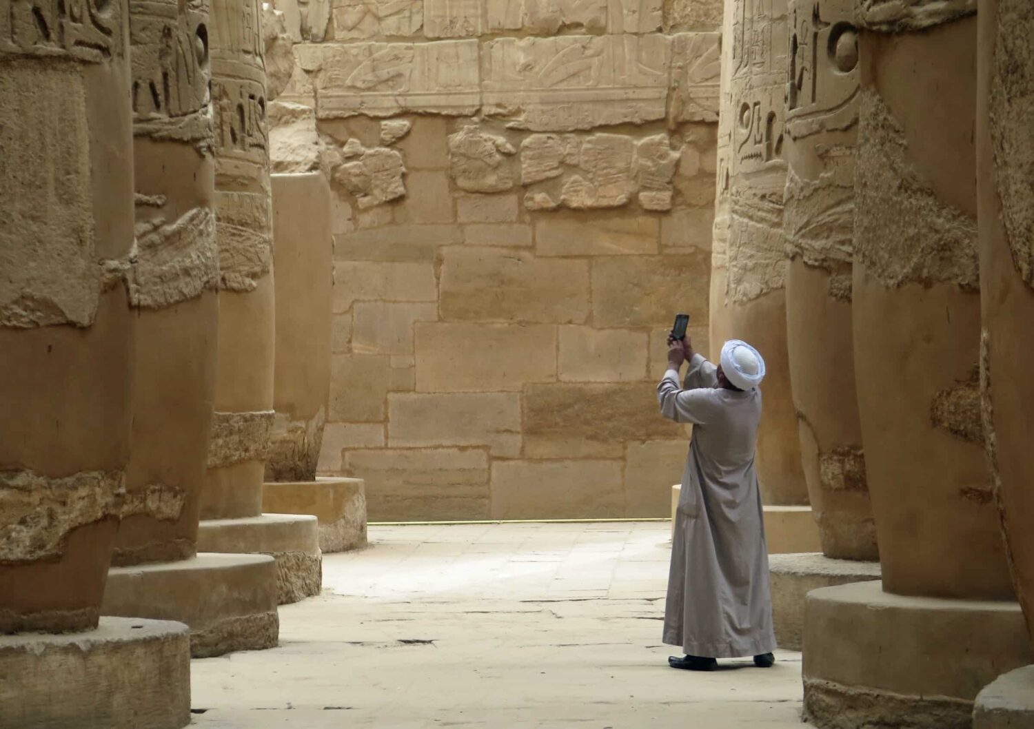 homem de costas com roupas típicas árabes tirando uma foto com o celular de um templo em Luxor, com várias colunas de pedra, que ilustra o post de chip celular Egito