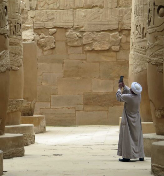 homem de costas com roupas típicas árabes tirando uma foto com o celular de um templo em Luxor, com várias colunas de pedra, que ilustra o post de chip celular Egito