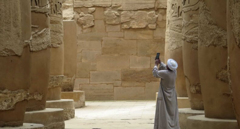 homem de costas com roupas típicas árabes tirando uma foto com o celular de um templo em Luxor, com várias colunas de pedra, que ilustra o post de chip celular Egito