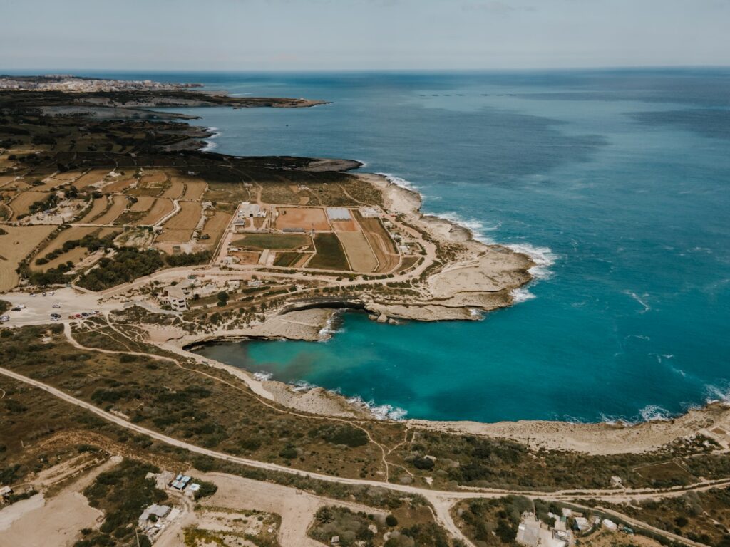 Vista aérea da região de Marsaxlokk, em Malta, com um mar azul turquesa e região costeira com campos verdes, estradas de terra e bastante vegetação
