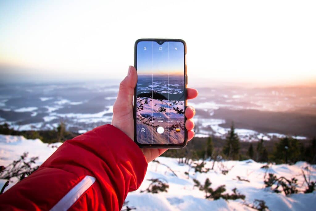 Uma mão masculina branca segurando um celular na vertical enquanto tira uma fotografia de montanhas cobertas por neve