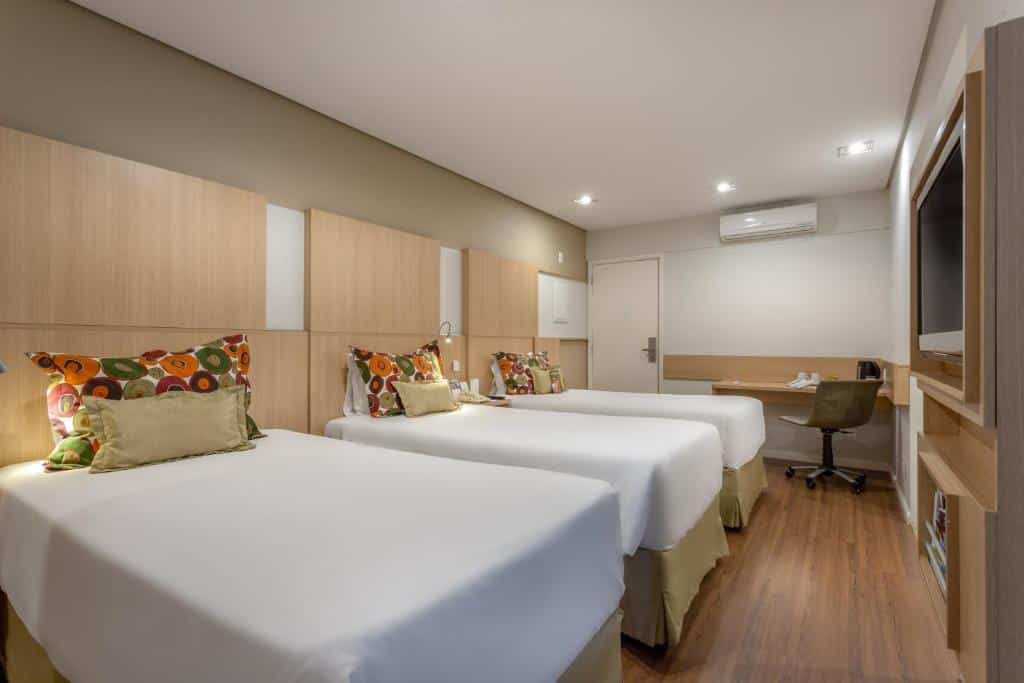 Quarto no Mercure Sao Paulo Naçoes Unidas com três camas de solteiro, um ar-condicionado, televisão, uma mesa de escritório com uma cadeira giratória, o chão é de madeira, para representar hotéis em Lollapalooza