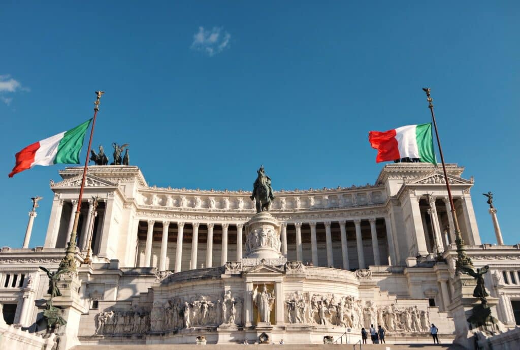 Uma construção histórica em Roma, com uma arquitetura colonial, com muitas colunas erguendo a construção e, na frente, há muitas esculturas, incluindo de um homem em um cavalo e, dos dois lados do local, há bandeiras da Itália hasteadas
