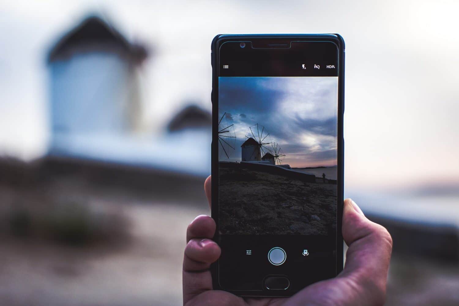 Uma mão segurando o celular, com a câmera ligada, mostrando a imagem dos moinhos de vento de Mykonos num fim de tarde