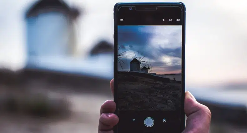 Uma mão segurando o celular, com a câmera ligada, mostrando a imagem dos moinhos de vento de Mykonos num fim de tarde