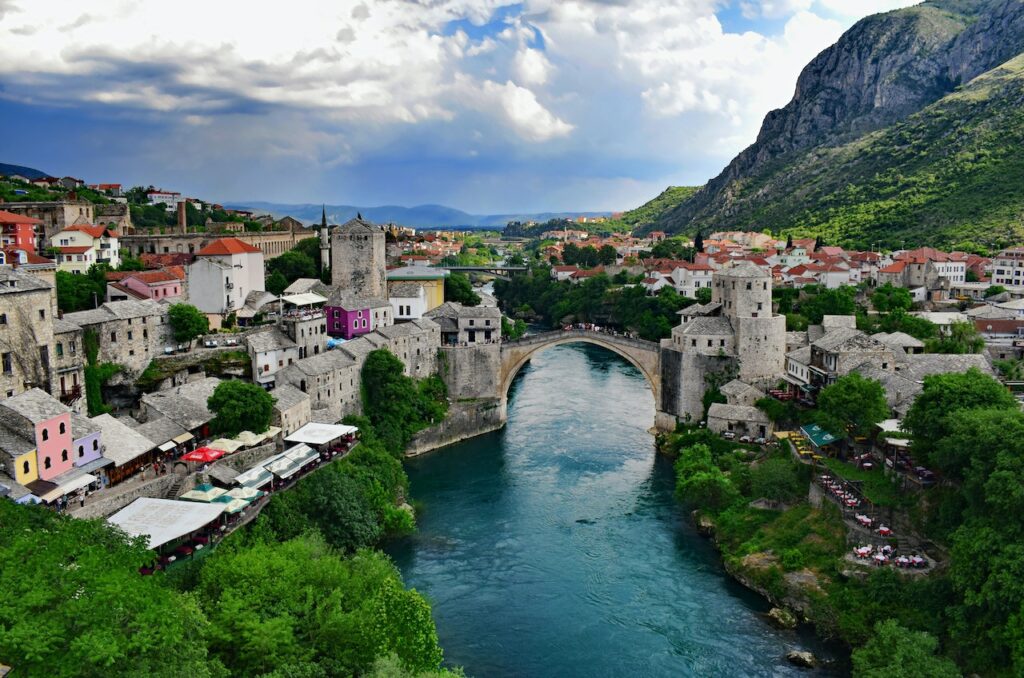 Rio azulado passando no meio de Mostar, cidade da Bósnia e Herzegovina, com árvores verdes e casas aos lados, e uma ponte no meio ligando duas regiões