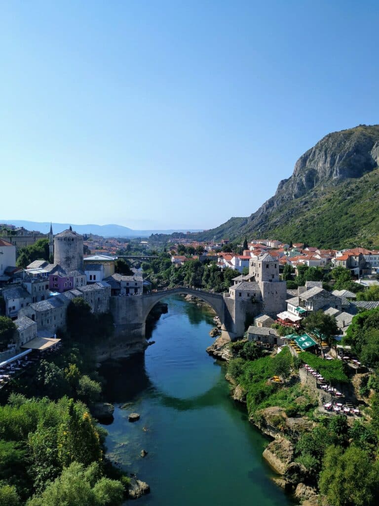Ponte de concreto acima de um rio para ilustrar o seguro viagem Bósnia e Herzegovina. Ao redor há casas e construções, além de árvores e vegetação. - Foto: Joshual Balzani via Unsplash