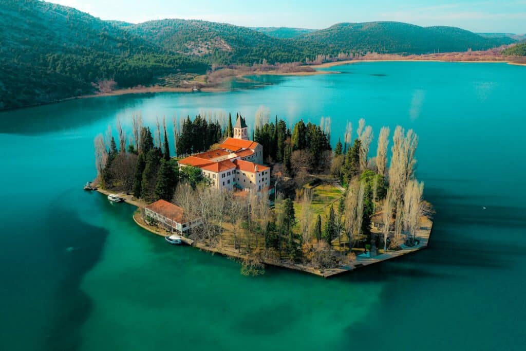 Pequena ilha, rodeada de um mar azul cristalino, com árvores cercando-a, além de dois prédios, sendo um o mosteiro de Visovac