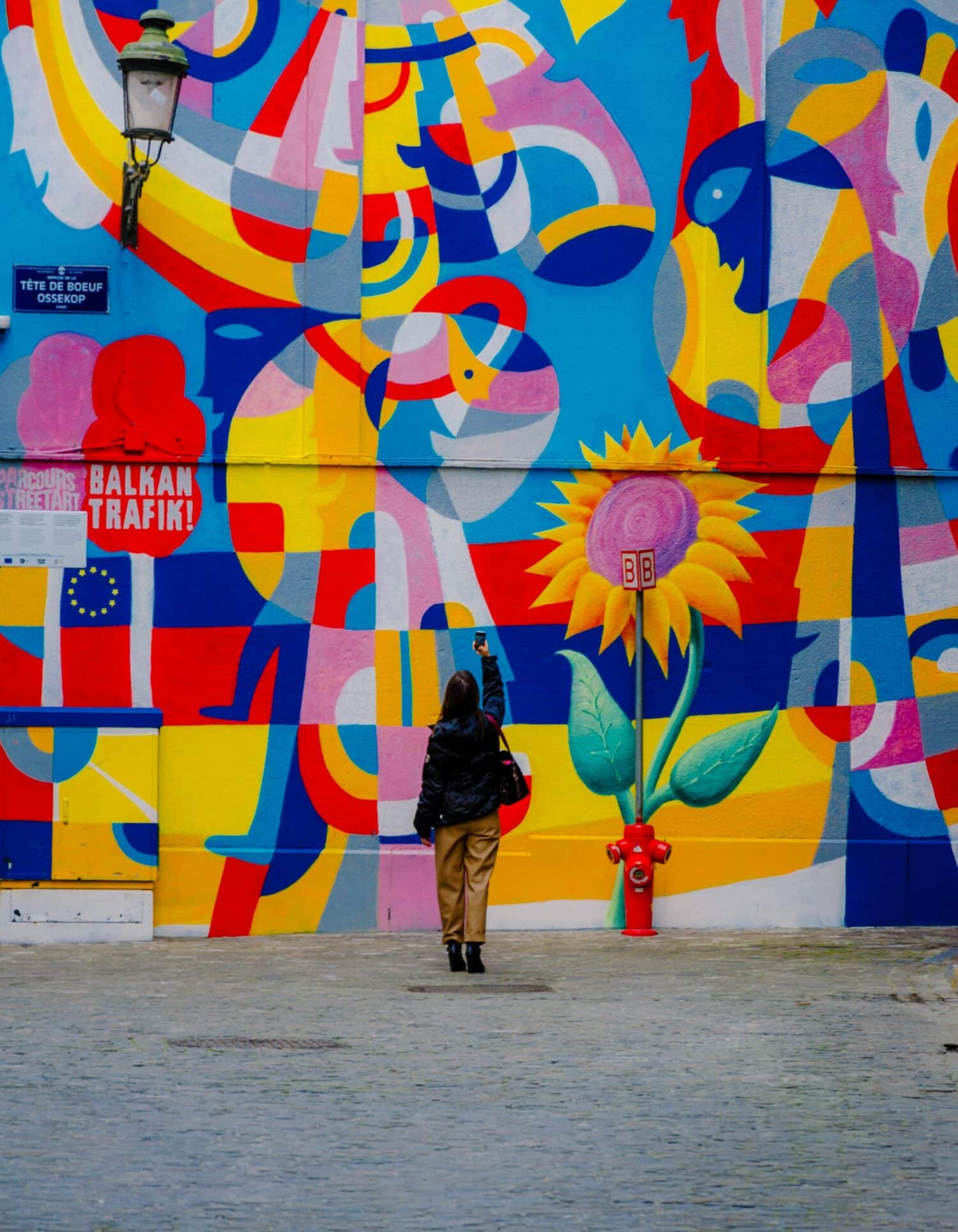 mulher de costas e roupas de frio segurando um celular acima da cabeça para fotografar um mural colorido com pessoas e flores abstratas em Bruxelas, na Bélgica