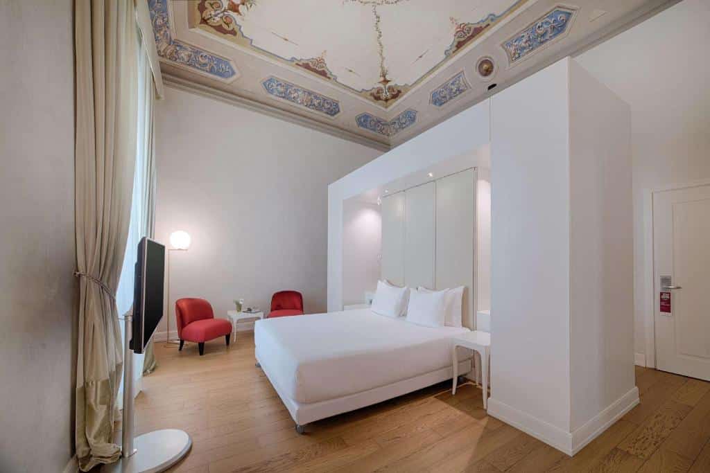 Quarto intimista do NH Collection Firenze Porta Rossa, com cama e paredes brancas, teto com pinturas, TV e duas poltronas vermelhas