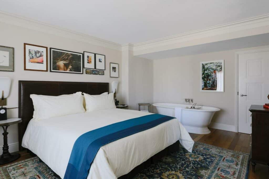 Quarto no NoMad Las Vegas com uma cama de casal do lado esquerdo, com alguns quadros sob a cama, o chão é de madeira e há um tapete em tons de azul, do lado direito tem uma banheira oval com toalhas penduradas