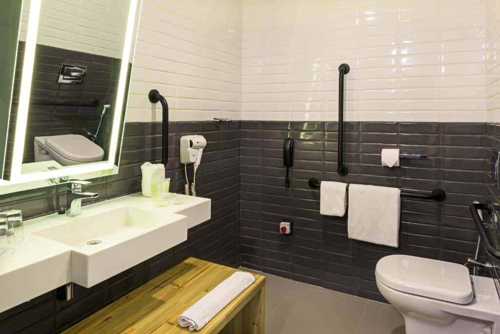 Banheiro amplo adaptado do Novotel Sao Paulo Morumbi com um vaso sanitário com barras de apoio, pia mais baixa com barras de apoio e um banco para se sentar