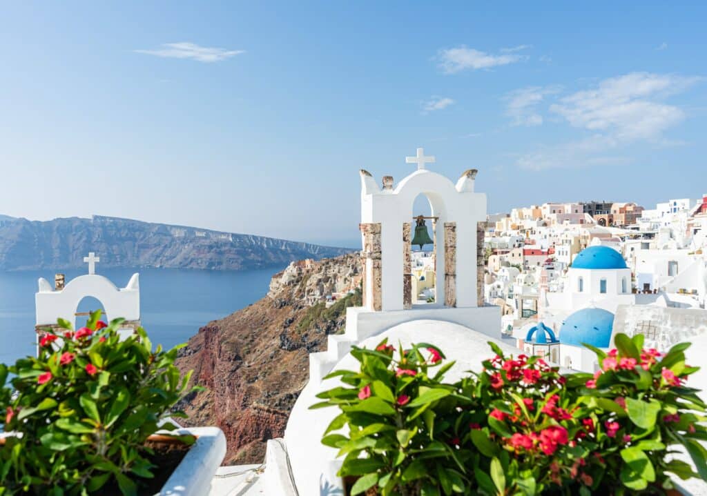 vista de cima da cidade de Oia, com flores rosas e casinhas brancas, com um sino azulado e vista da orla com o mar bem azul de fundo, que ilustra o post de chip celular Grécia