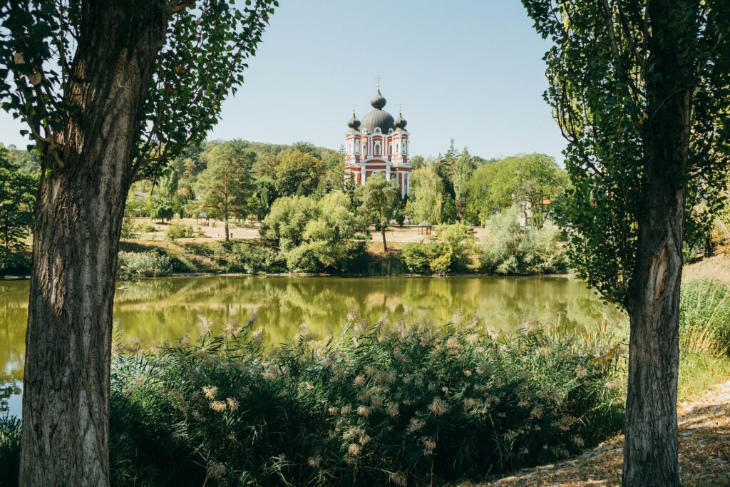 Um amplo lago em tom de verde que fica em frente uma monastério em Moldávia, o monastério é pequeno mas bem alto, é pintado de branco e vermelho, com janelas arredondadas e no topo das três torres há uma abóboda em tom de verde, ao redor da construção há muito verde e árvores