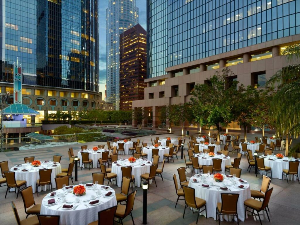 Terraço do Omni Los Angeles Hotel California Plaza com muitas mesas redondas ao ar livre e cadeiras estofadas em bege, há também árvores e iluminação indireta no local