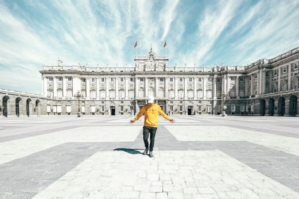 Pessoa com boné branco, blusa de manga longa amarela e calça jeans preta, com os braços abertos, olhando para baixo, em frente ao Palácio Real de Madri, num dia ensolarado, com o céu azul e algumas nuvens brancas