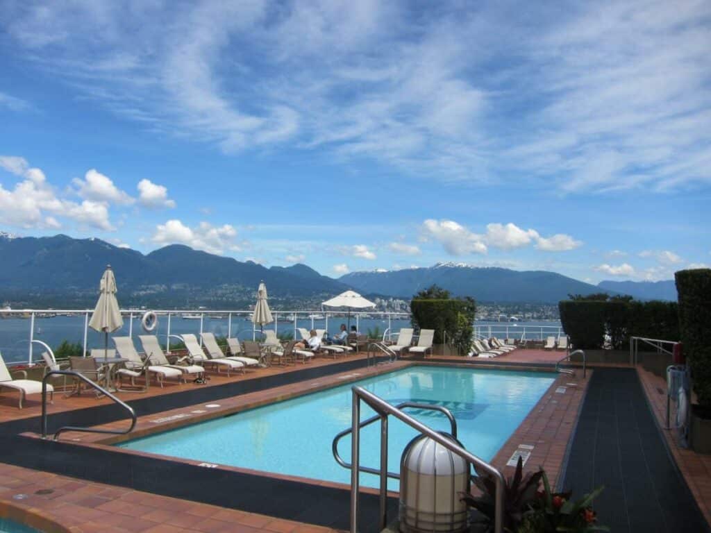 Piscina na cobertura do Pan Pacific Vancouver com vista para a baía e, ao redor, há um deck com espreguiçadeiras brancas com guarda-sóis