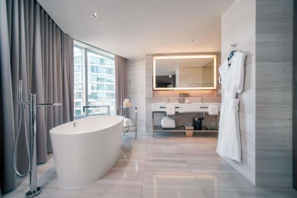 Banheiro espaçoso do Paradox Hotel Vancouver com uma banheira oval ao lado de um janela com cortinas, o chão é porcelanato, há um pequeno cabide com roupões pendurados, ao fundo do local, há uma pia ampla com duas torneiras e uma espelho com iluminação ao redor, para representar hotéis em Vancouver