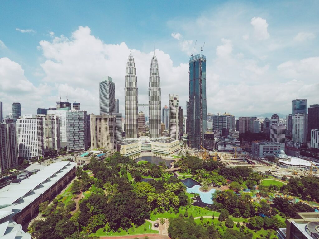 Parque com vasta vegetação verde ao centro, um lago e construções ao entorno, como prédios e arranha céus modernos em um dia azul com algumas nuvens para representar o seguro viagem Kuala Lumpur.
