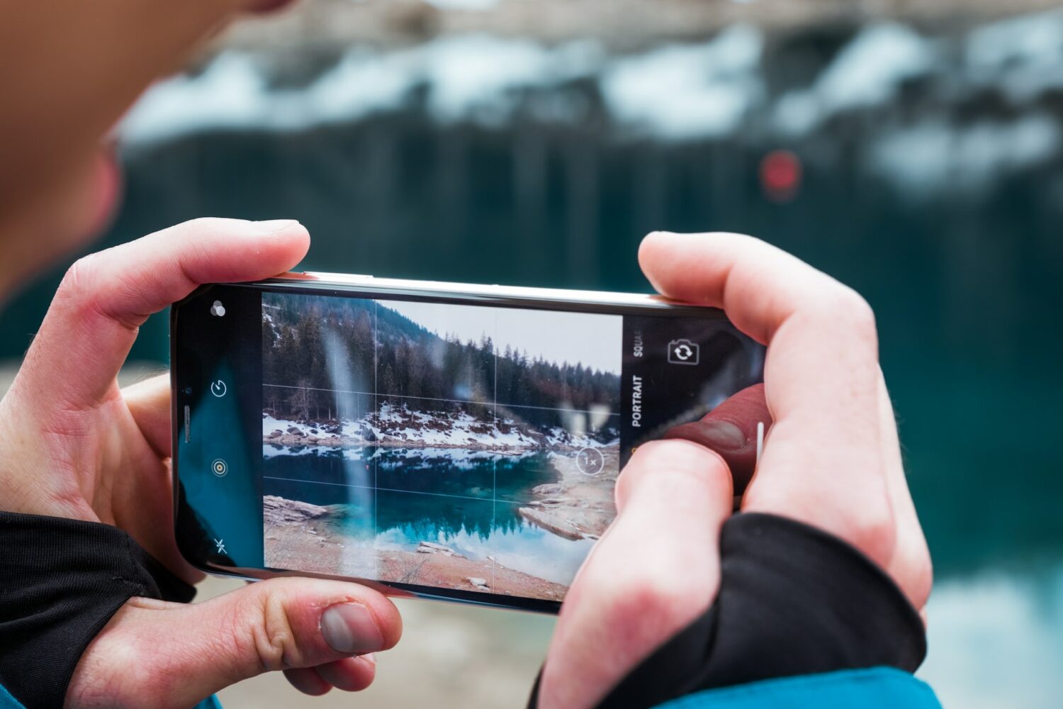 Duas mãos masculinas brancas segurando um celular na horizontal para fotografar um lago de azul azuis cercado por árvores