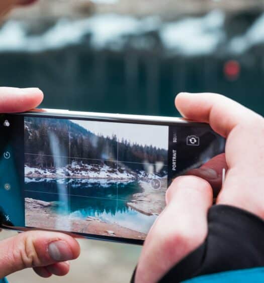 Duas mãos masculinas brancas segurando um celular na horizontal para fotografar um lago de azul azuis cercado por árvores