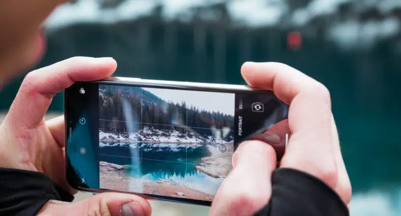 Duas mãos masculinas brancas segurando um celular na horizontal para fotografar um lago de azul azuis cercado por árvores