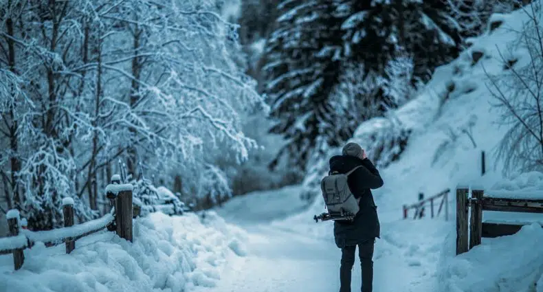 Pessoa com uma mochila nas costas etirando foto de algo em sua frente. Ao redor, há uma cerca e algumas árvores covertas de neve, assim como o chão. - Foto: Armin Djuhic via Unsplash