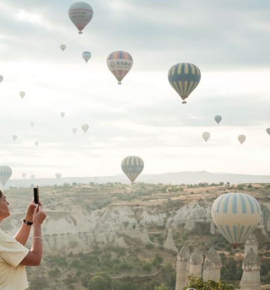 Mulher branca e loira de perfil tirando foto dos balões que sobrevoam o céu da Capadócia. - Foto: Christina Kuklina via Pexels