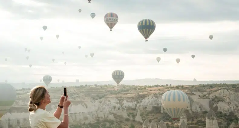 Mulher branca e loira de perfil tirando foto dos balões que sobrevoam o céu da Capadócia. - Foto: Christina Kuklina via Pexels