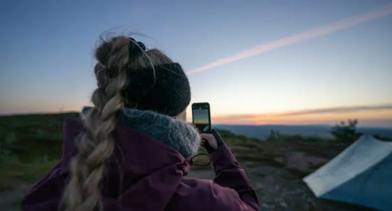 Mulher branca e com trança loira tirando foto do pôr do sol. Ela usa um moletom roxo e à direita podemos ver uma barraca branca. - Foto: Hendrik Morkel via Unsplash