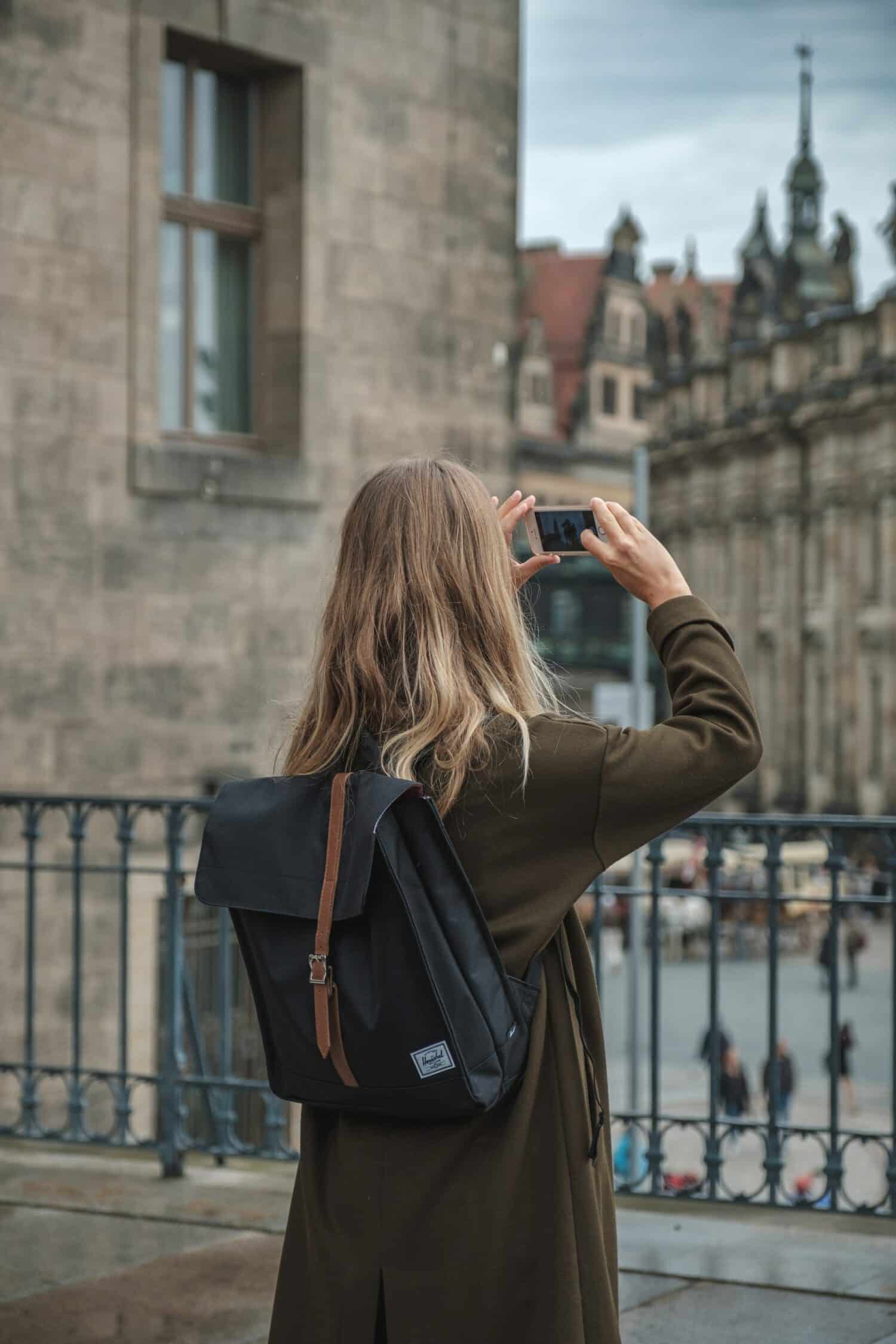 Mulher loira com casaco cinza de costas tirando foto do centro histórico de Dresden, que possui arquitetura clássica. - Foto: Jan Baborák via Unsplash