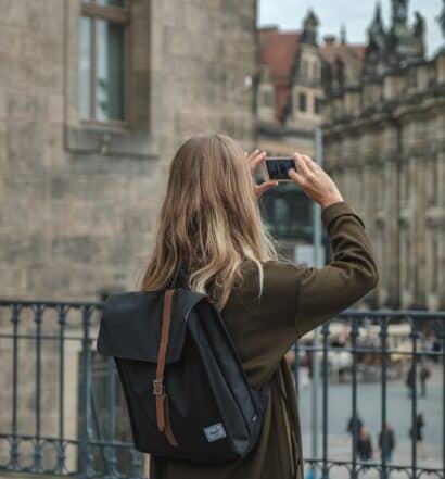 Mulher loira com casaco cinza de costas tirando foto do centro histórico de Dresden, que possui arquitetura clássica. - Foto: Jan Baborák via Unsplash