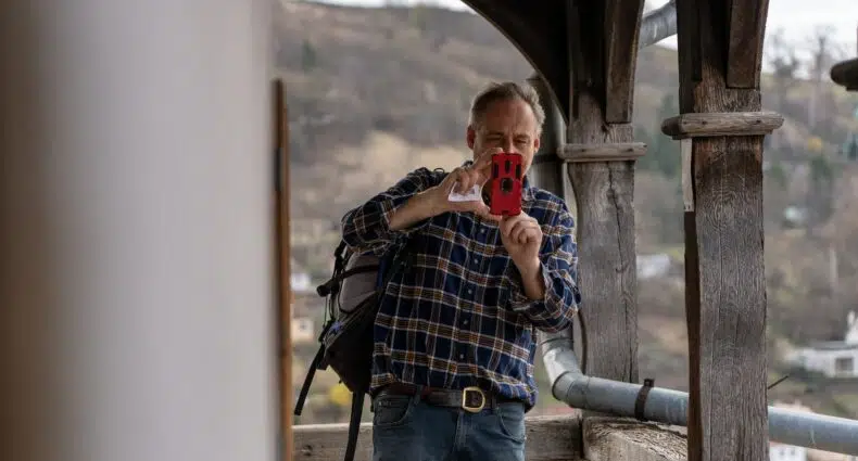 Homem de camisa de flanela azul marinho em meio a ruínas de uma construção apontando o celular para a frente. - Foto: Jonny Gios via Unsplash