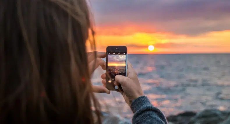 Mulher de cabelos castanhos tirando foto do por do sol na praia. - Foto: Mantas Hesthaven via Unsplash