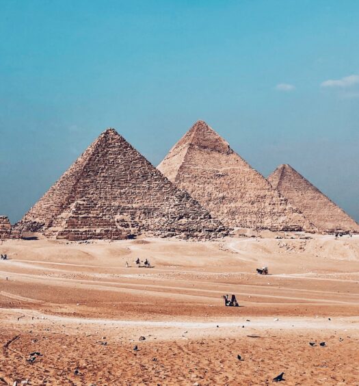 as três Pirâmides de Gizé, no Egito, em diferentes tamanho em meio ao deserto em tons de amarelo e alaranjado e ceu azul