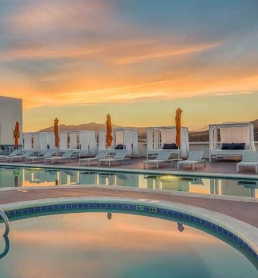 Uma piscina do Hampton Inn & Suites by Hilton Los Cabos com cabanas e guarda-sóis à beira da piscina ao pôr do sol, em meio a colinas distantes e um céu parcialmente nublado. Representa hotéis em Los Cabos.