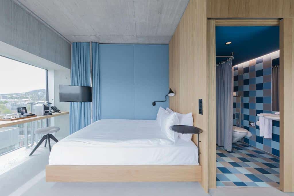 Quarto acessível do Placid Hotel Design, em Zurique, com cama de casal, TV, parede de vidro mostrando a cidade, balcão com utensílios para fazer café e banheiro adaptado com barras de apoio